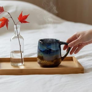 May include: A wooden tray on a bed holds a clear glass bottle with red leaves and a steaming blue and brown ceramic mug. The mug is being held by a hand. The scene suggests a cozy, relaxing morning.
