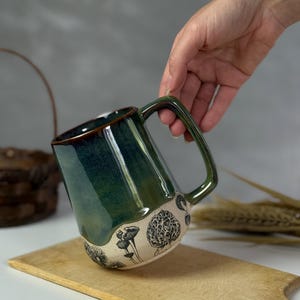 May include: A handmade ceramic mug with a dark green glaze and a brown rim. The bottom of the mug features a cream-colored band with black mushroom illustrations. The mug is held over a wooden cutting board.