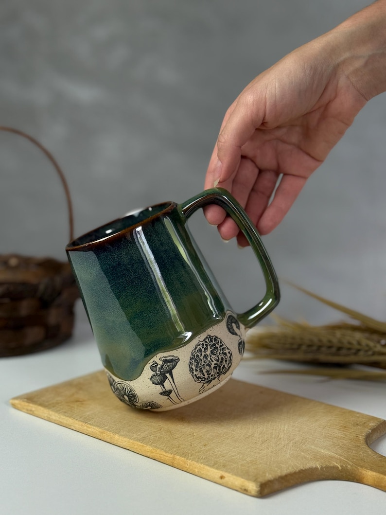 May include: A handmade ceramic mug with a dark green glaze and a brown rim. The bottom of the mug features a cream-colored band with black mushroom illustrations. The mug is held over a wooden cutting board.