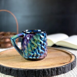 Pu&ograve; includere: Una piccola tazza in ceramica strutturata con manico, nei toni del blu, verde e viola. La tazza &egrave; appoggiata su un vassoio rotondo di legno con un bordo scuro e strutturato. Sullo sfondo &egrave; visibile un libro aperto.