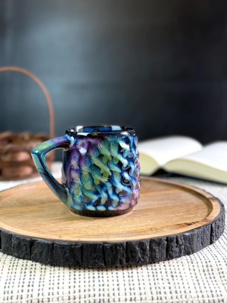 Pu&ograve; includere: Una piccola tazza in ceramica strutturata con manico, nei toni del blu, verde e viola. La tazza &egrave; appoggiata su un vassoio rotondo di legno con un bordo scuro e strutturato. Sullo sfondo &egrave; visibile un libro aperto.