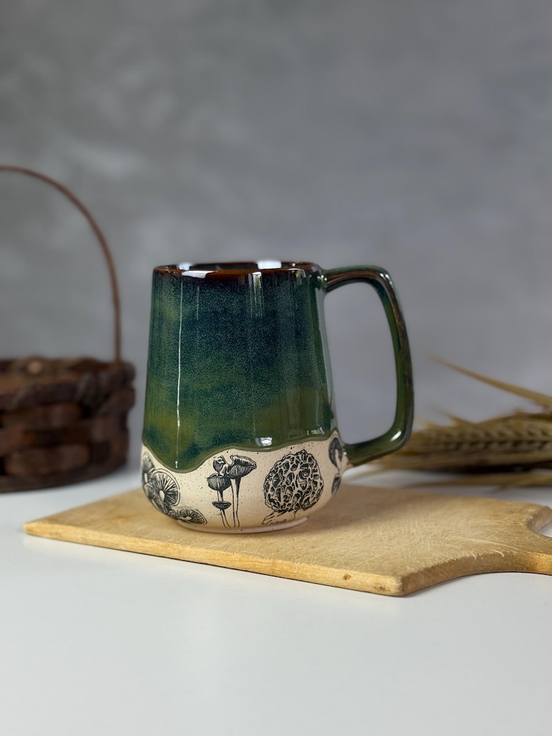 May include: A handmade ceramic mug with a dark green glaze and a brown rim. The lower portion of the mug features black and white illustrations of various mushrooms. The mug is set on a wooden cutting board.