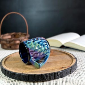 Pu&ograve; includere: Una tazza in ceramica con una glassa iridescente strutturata nei toni del blu, verde e viola. La tazza &egrave; appoggiata su un vassoio di legno con un bordo scuro e strutturato. Un cestino intrecciato e un libro aperto sono sullo sfondo.