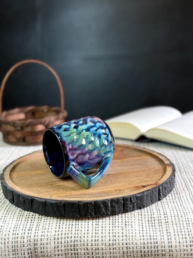 Pu&ograve; includere: Una tazza in ceramica con una glassa iridescente strutturata nei toni del blu, verde e viola. La tazza &egrave; appoggiata su un vassoio di legno con un bordo scuro e strutturato. Un cestino intrecciato e un libro aperto sono sullo sfondo.