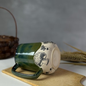 May include: A ceramic mug with a green glazed top and handle, and a cream-colored body decorated with black mushroom illustrations. The mug is resting on a wooden cutting board.
