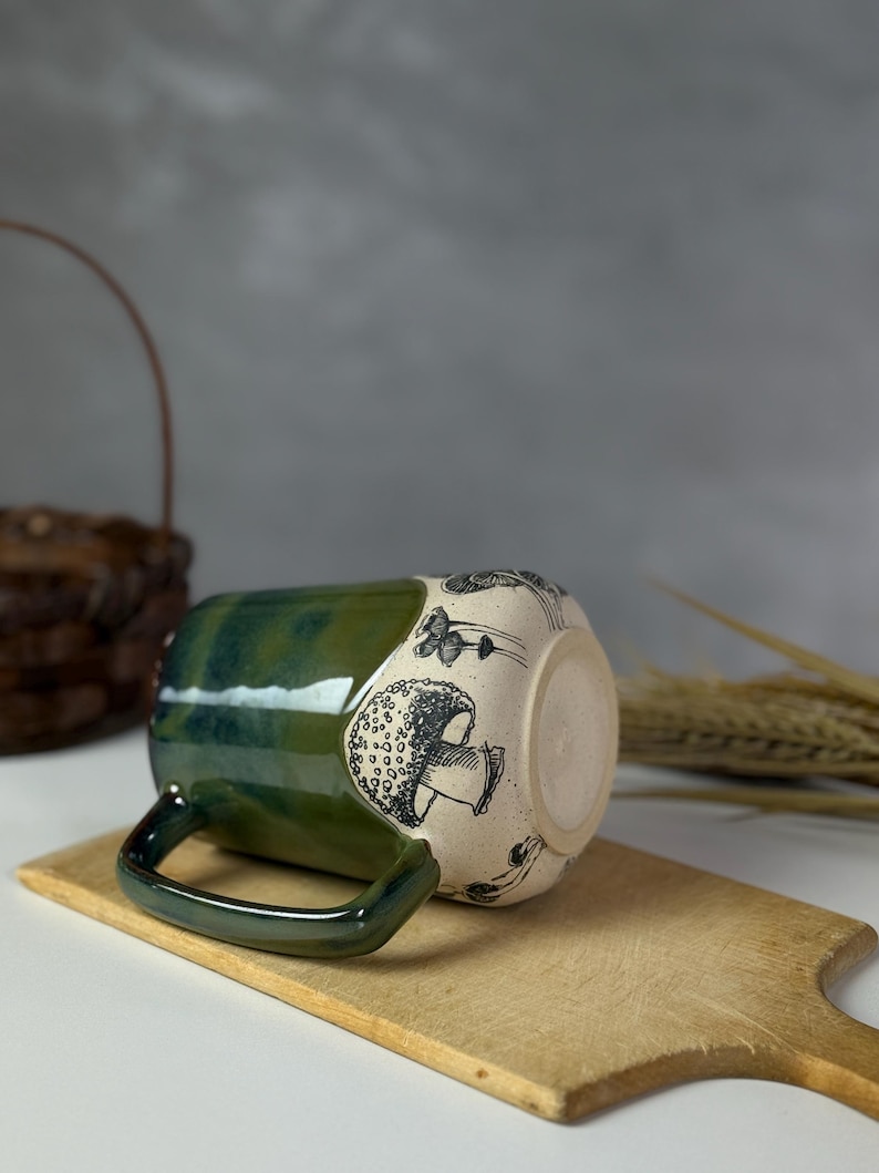 May include: A ceramic mug with a green glazed top and handle, and a cream-colored body decorated with black mushroom illustrations. The mug is resting on a wooden cutting board.
