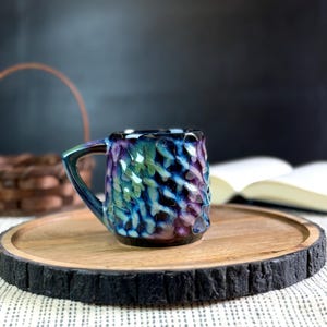Pu&ograve; includere: Una piccola tazza in ceramica fatta a mano con una superficie strutturata e una glassa colorata nelle tonalit&agrave; del blu, viola e verde. La tazza &egrave; appoggiata su un vassoio rotondo di legno con un bordo scuro e strutturato. Un libro e un cestino sono sullo sfondo.