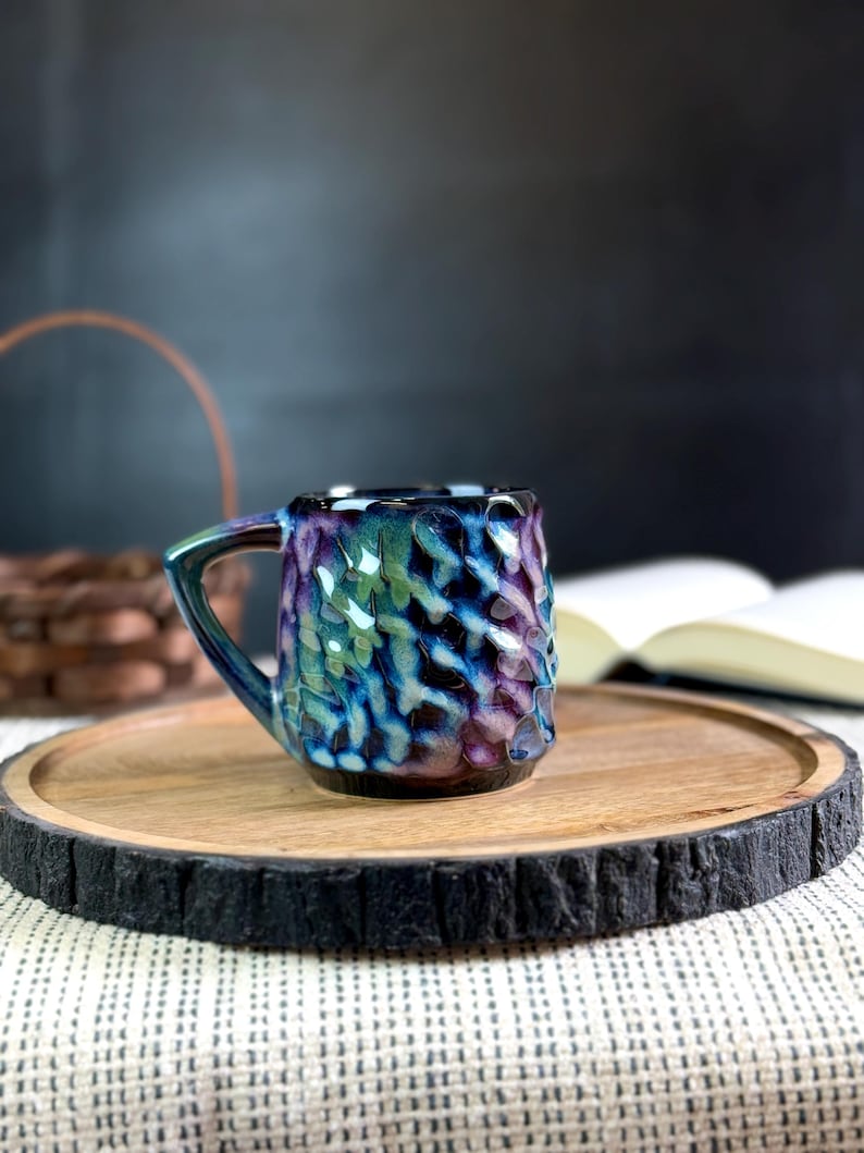 Pu&ograve; includere: Una piccola tazza in ceramica fatta a mano con una superficie strutturata e una glassa colorata nelle tonalit&agrave; del blu, viola e verde. La tazza &egrave; appoggiata su un vassoio rotondo di legno con un bordo scuro e strutturato. Un libro e un cestino sono sullo sfondo.