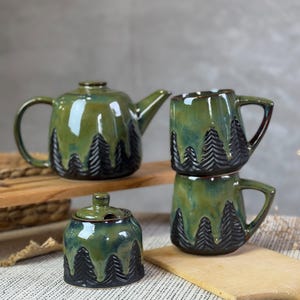 May include: A green ceramic tea set with a forest design. The set includes a teapot, two mugs, and a sugar bowl. The forest design is black and covers the bottom half of each piece. The set is on a wooden surface.