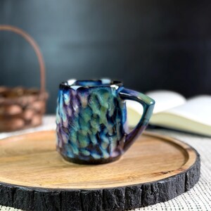 Pu&ograve; includere: Una piccola tazza in ceramica strutturata con una glassa blu, verde e viola. La tazza ha un manico e poggia su un vassoio rotondo di legno con un bordo scuro e strutturato. Un cestino e un libro sono sullo sfondo.