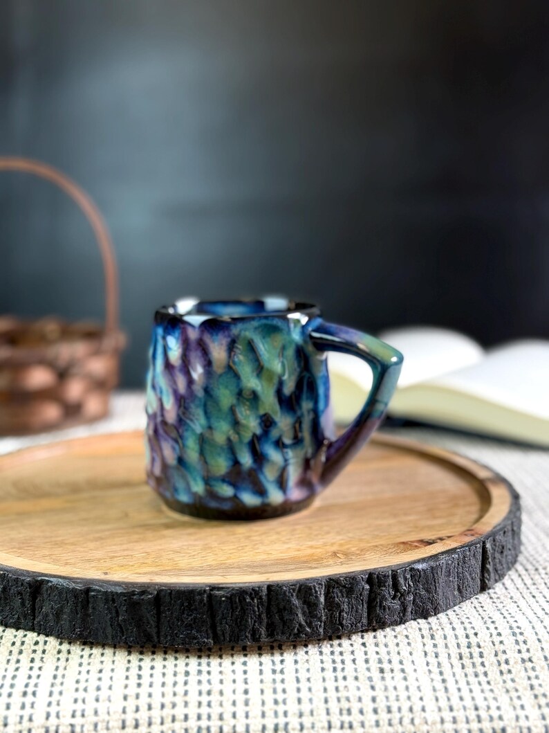 Pu&ograve; includere: Una piccola tazza in ceramica strutturata con una glassa blu, verde e viola. La tazza ha un manico e poggia su un vassoio rotondo di legno con un bordo scuro e strutturato. Un cestino e un libro sono sullo sfondo.