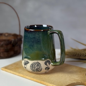 May include: A handmade ceramic mug with a green and blue glaze. The bottom of the mug features black and white mushroom illustrations. The mug has a large handle and is sitting on a wooden cutting board.