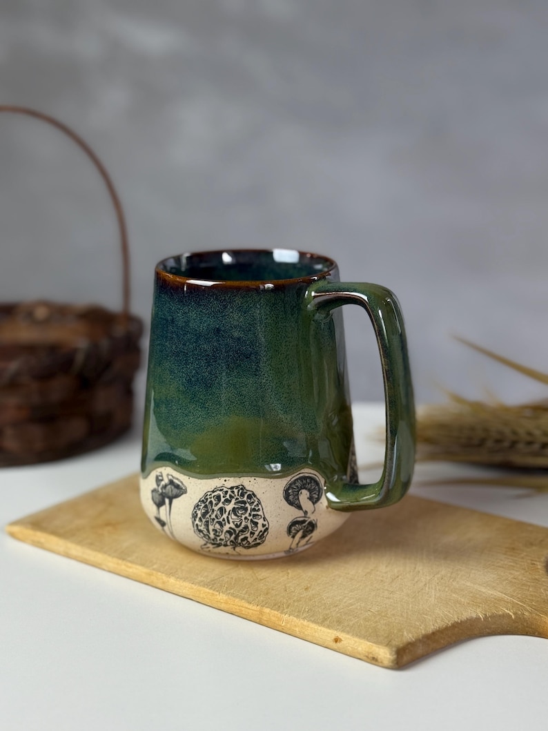 May include: A handmade ceramic mug with a green and blue glaze. The bottom of the mug features black and white mushroom illustrations. The mug has a large handle and is sitting on a wooden cutting board.