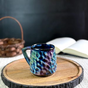 Pu&ograve; includere: Una tazza in ceramica strutturata con smalto blu, verde e viola &egrave; appoggiata su un vassoio di legno. La tazza ha un manico e una superficie unica e increspata. Il vassoio ha un bordo naturale ed &egrave; posto su una superficie a motivi.