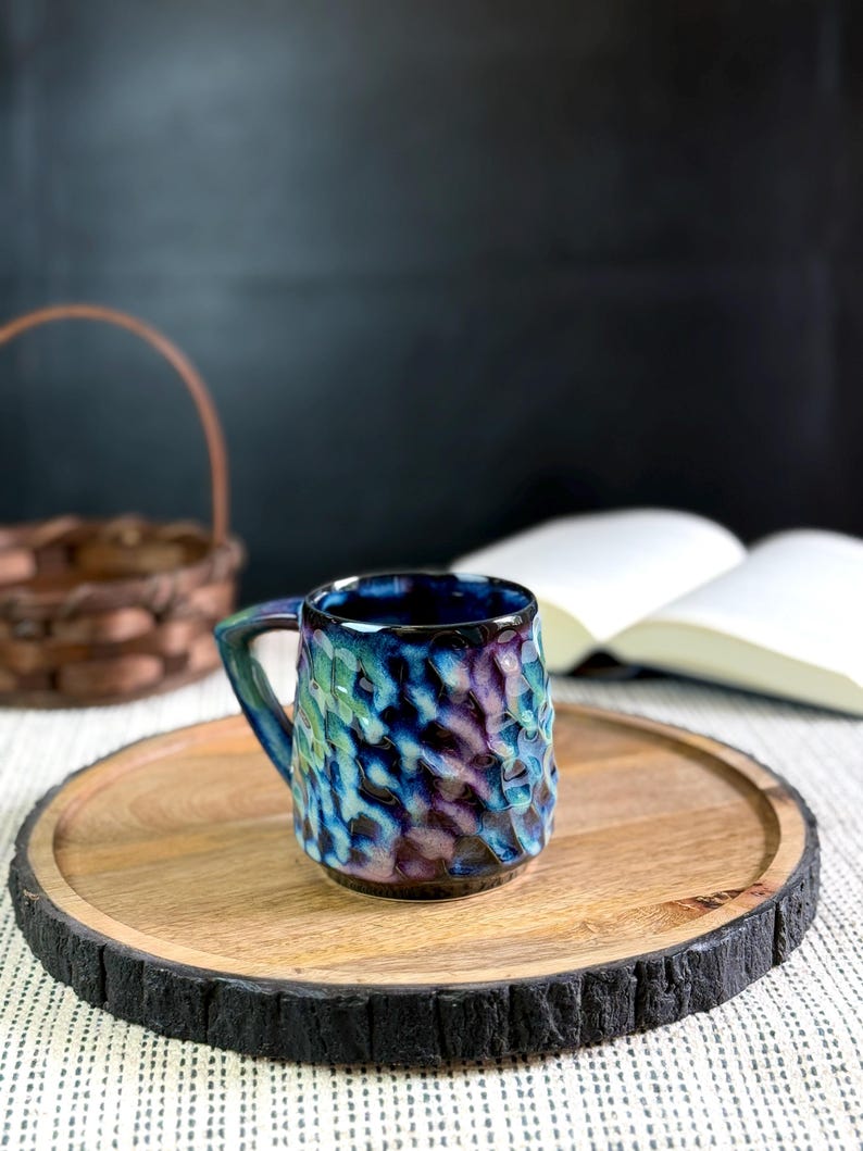 Pu&ograve; includere: Una tazza in ceramica strutturata con smalto blu, verde e viola &egrave; appoggiata su un vassoio di legno. La tazza ha un manico e una superficie unica e increspata. Il vassoio ha un bordo naturale ed &egrave; posto su una superficie a motivi.