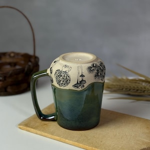 May include: A ceramic mug with a green and cream glaze. The mug features a dark green handle and a band of black mushroom illustrations around the rim. The mug is resting on a wooden cutting board.