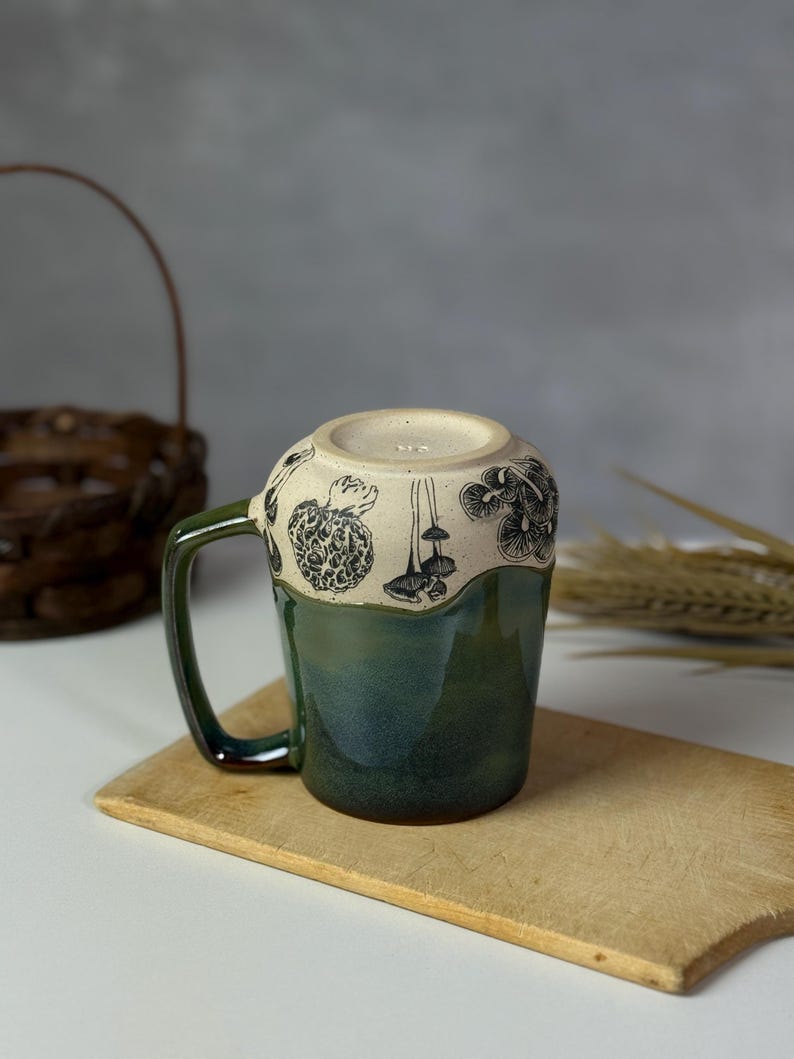 May include: A ceramic mug with a green and cream glaze. The mug features a dark green handle and a band of black mushroom illustrations around the rim. The mug is resting on a wooden cutting board.