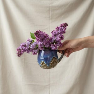 Peut inclure: Une tasse en céramique bleue avec une anse, remplie de fleurs de lilas mauves. La tasse a un extérieur texturé, marron et bleu. Les fleurs sont d'un violet vif, avec des feuilles vertes. La tasse est tenue sur un fond neutre.