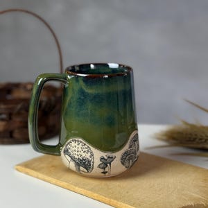 May include: A ceramic mug with a green and blue glaze, featuring black and white mushroom illustrations around the base. The mug has a large handle and sits on a wooden cutting board.