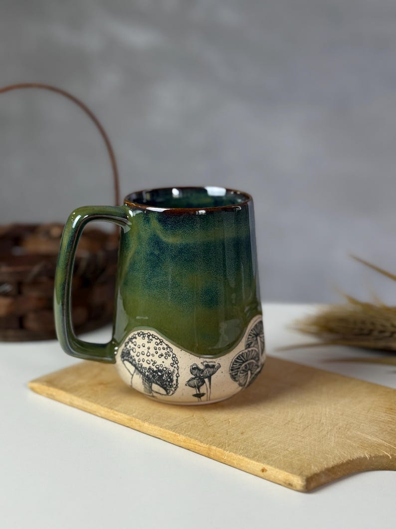 May include: A ceramic mug with a green and blue glaze, featuring black and white mushroom illustrations around the base. The mug has a large handle and sits on a wooden cutting board.