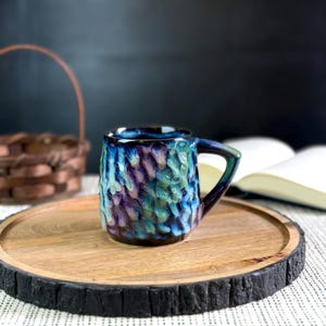 Pu&ograve; includere: Una tazza in ceramica fatta a mano con una superficie strutturata e un manico blu scuro. La tazza presenta una miscela di colori blu, viola e verde. &Egrave; appoggiata su un vassoio di legno con un bordo scuro e strutturato.
