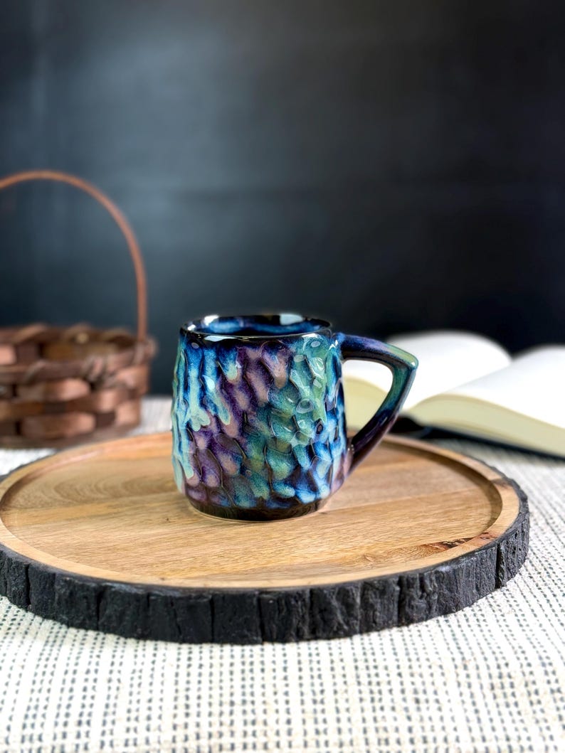 Pu&ograve; includere: Una tazza in ceramica fatta a mano con una superficie strutturata e un manico blu scuro. La tazza presenta una miscela di colori blu, viola e verde. &Egrave; appoggiata su un vassoio di legno con un bordo scuro e strutturato.