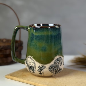 May include: A ceramic mug with a green and blue glaze. The mug features a black and white mushroom design around the base. The handle is a matching green color. The mug is sitting on a wooden cutting board.