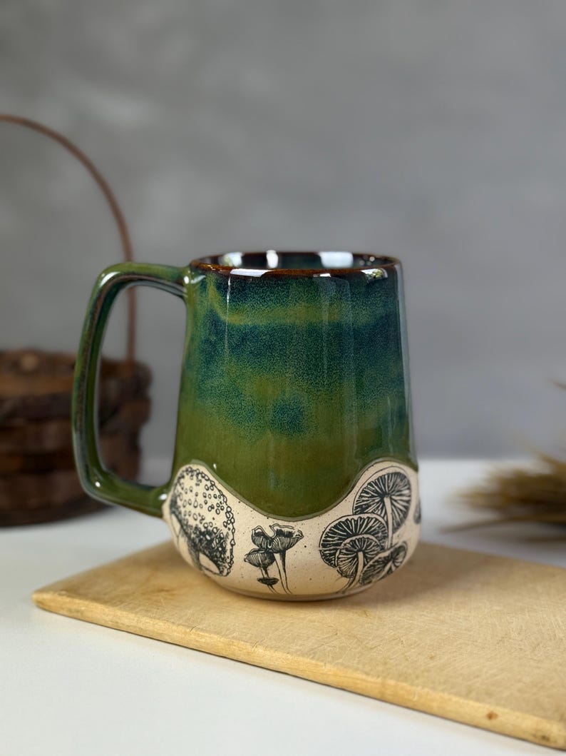 May include: A ceramic mug with a green and blue glaze. The mug features a black and white mushroom design around the base. The handle is a matching green color. The mug is sitting on a wooden cutting board.
