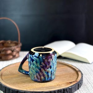 Pu&ograve; includere: Una tazza in ceramica con superficie strutturata e manico. La tazza presenta una miscela di colori blu, viola e verde. &Egrave; appoggiata su un vassoio di legno con bordo scuro e strutturato. Un libro e un cestino sono sullo sfondo.