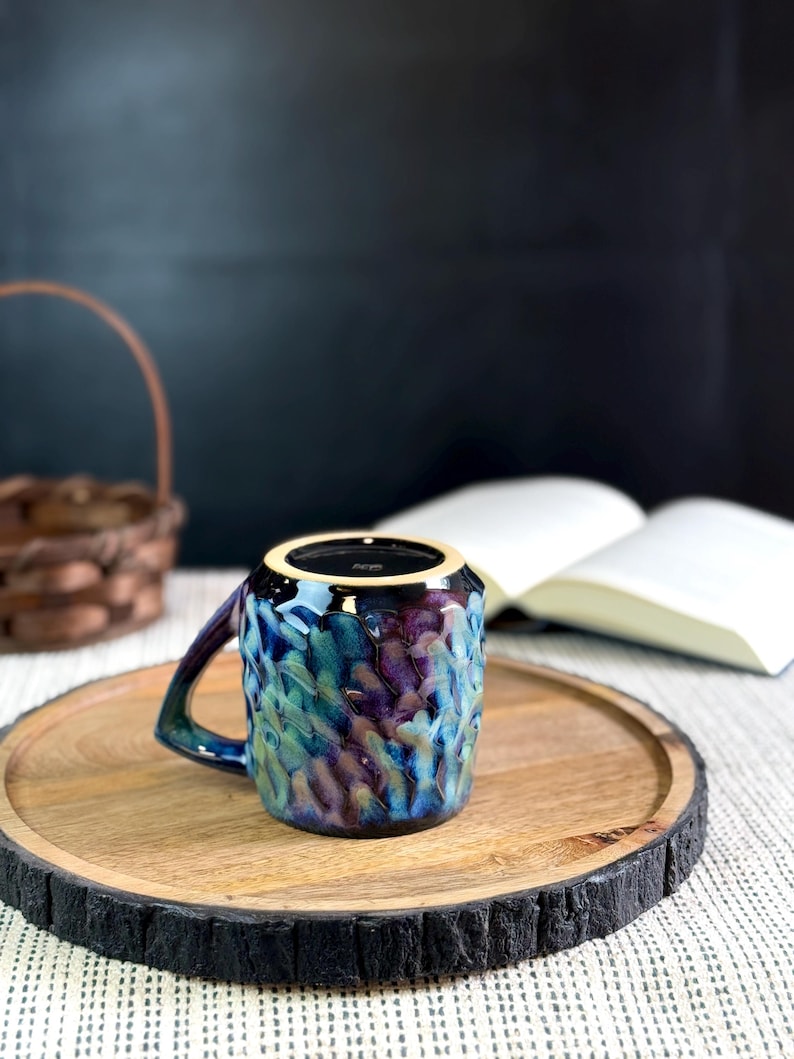Pu&ograve; includere: Una tazza in ceramica con superficie strutturata e manico. La tazza presenta una miscela di colori blu, viola e verde. &Egrave; appoggiata su un vassoio di legno con bordo scuro e strutturato. Un libro e un cestino sono sullo sfondo.