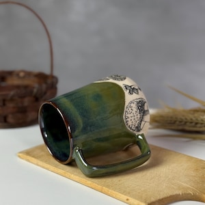May include: A ceramic mug with a green and brown glaze. The mug features a handle and a decorative design of butterflies and other shapes. The mug is sitting on a wooden cutting board.