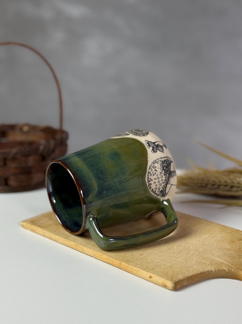 May include: A ceramic mug with a green and brown glaze. The mug features a handle and a decorative design of butterflies and other shapes. The mug is sitting on a wooden cutting board.