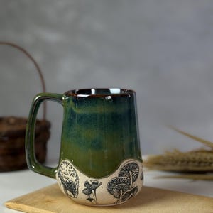 May include: A ceramic mug with a green and brown glaze. The mug features a black and white illustration of mushrooms around the base. The mug has a rectangular handle and sits on a wooden board.