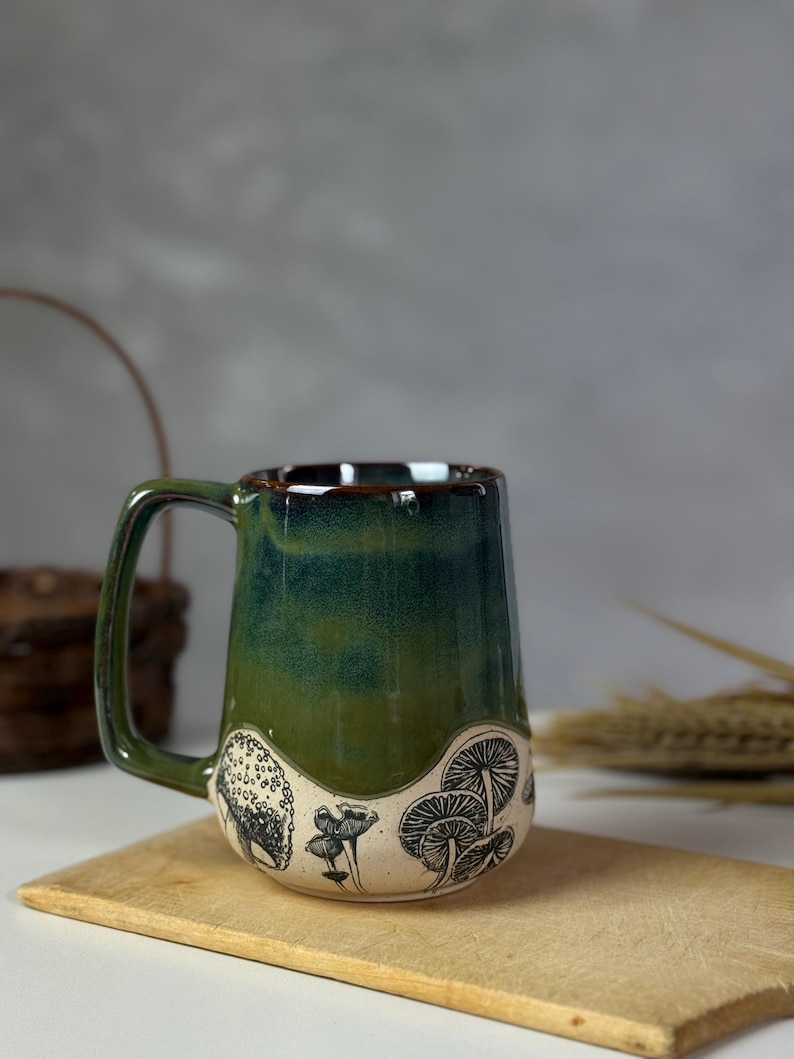 May include: A ceramic mug with a green and brown glaze. The mug features a black and white illustration of mushrooms around the base. The mug has a rectangular handle and sits on a wooden board.