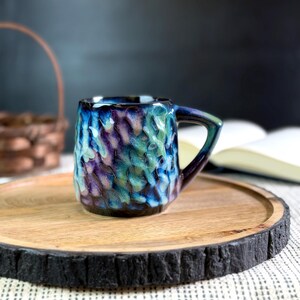 Pu&ograve; includere: Una piccola tazza in ceramica strutturata con manico. La tazza presenta una miscela di colori blu, verde acqua e viola. &Egrave; appoggiata su un vassoio rotondo di legno con un bordo scuro e strutturato. Un cestino e un libro aperto sono sfocati sullo sfondo.