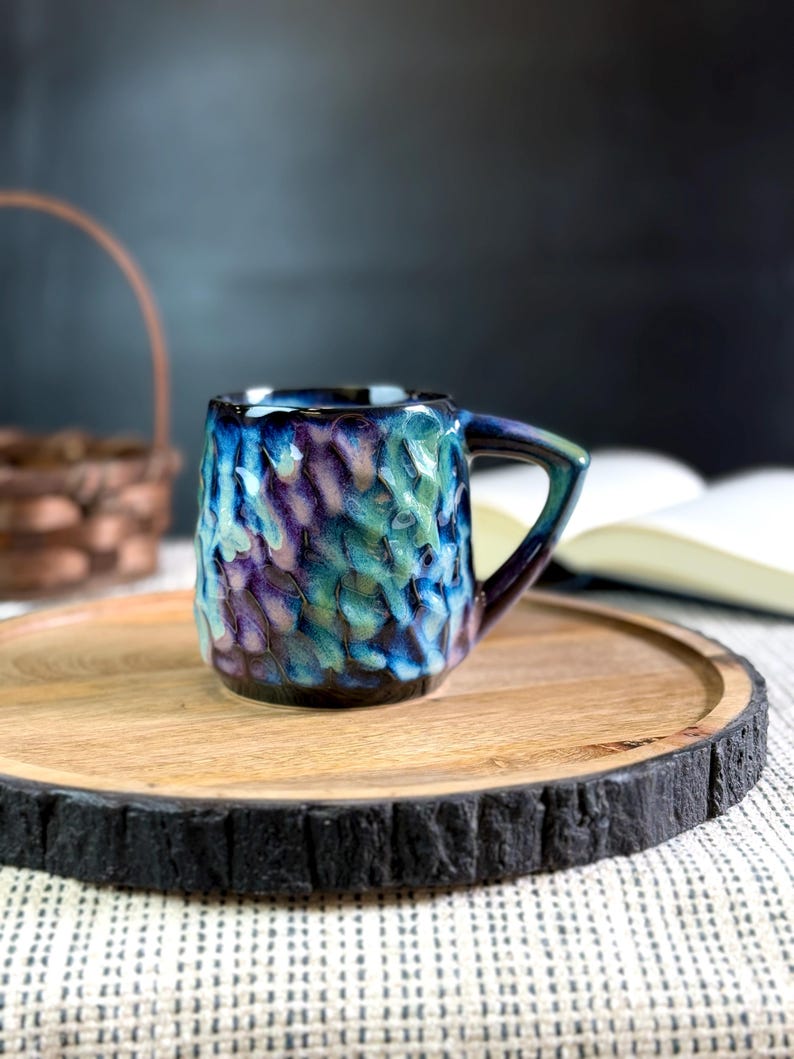 Pu&ograve; includere: Una piccola tazza in ceramica strutturata con manico. La tazza presenta una miscela di colori blu, verde acqua e viola. &Egrave; appoggiata su un vassoio rotondo di legno con un bordo scuro e strutturato. Un cestino e un libro aperto sono sfocati sullo sfondo.
