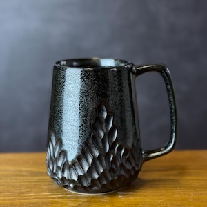 Op de afbeelding: Zwarte keramische mok met een getextureerd, gebeeldhouwd ontwerp op de onderste helft. De mok heeft een vierkant handvat en een glanzende afwerking. De mok staat op een houten oppervlak.