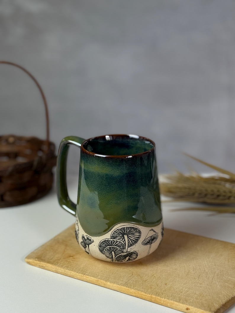 May include: A handmade ceramic mug with a dark green glaze and a black mushroom design. The mug has a sturdy handle and a wide opening. The mug is sitting on a wooden cutting board.