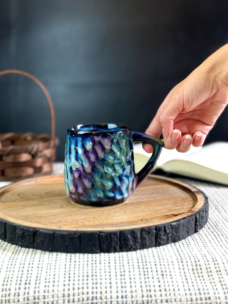 Pu&ograve; includere: Una tazza in ceramica fatta a mano con una superficie strutturata e una sfumatura di colore unica di blu, viola e verde. La tazza &egrave; appoggiata su un vassoio di legno con un bordo scuro e strutturato. Un cestino e un libro sono sullo sfondo.