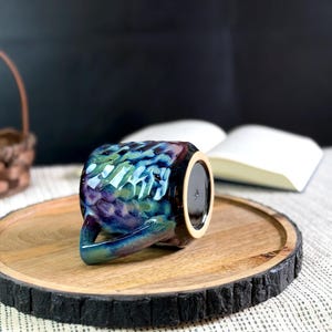 Pu&ograve; includere: Una tazza in ceramica con una glassa iridescente strutturata nei toni del blu, verde e viola. La tazza &egrave; appoggiata su un vassoio di legno con un bordo scuro e strutturato. Un libro &egrave; aperto sullo sfondo.