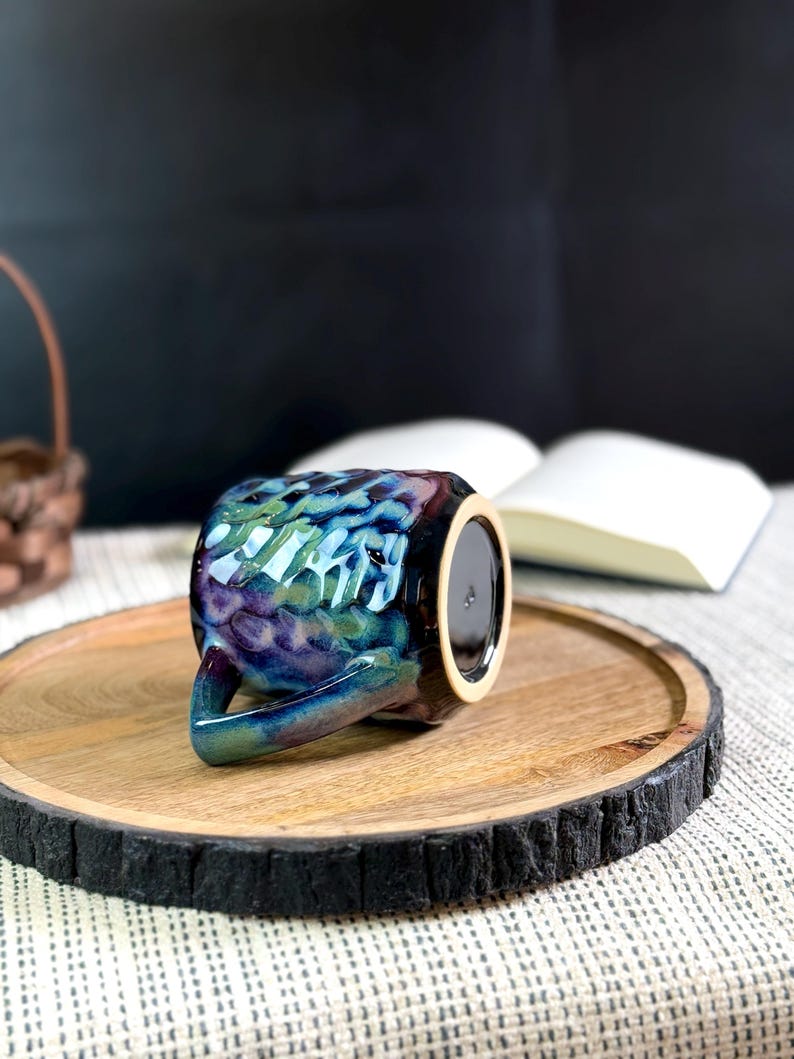 Pu&ograve; includere: Una tazza in ceramica con una glassa iridescente strutturata nei toni del blu, verde e viola. La tazza &egrave; appoggiata su un vassoio di legno con un bordo scuro e strutturato. Un libro &egrave; aperto sullo sfondo.