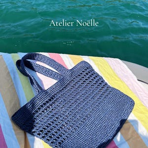 May include: A dark blue crochet tote bag with two handles and an open-weave pattern on the upper section. The bag features a solid base and is positioned on a striped fabric. The text Atelier Noëlle is visible in the upper center of the image.