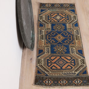 Puede incluir: Una pequeña alfombra rectangular con un patrón geométrico en tonos azules, naranjas y marrones. La alfombra presenta un campo azul central con formas geométricas repetidas y bordes. La alfombra está colocada sobre un suelo de madera clara.