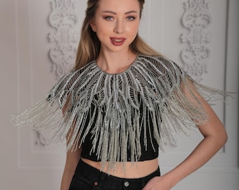 May include: A silver beaded collar necklace with fringe detailing. The necklace is worn by a person with long blonde hair and is styled with a black crop top and black jeans.