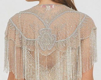 May include: A beaded shawl with a fringe border. The shawl is made of a sheer fabric and has a floral design. The shawl is a light beige color.