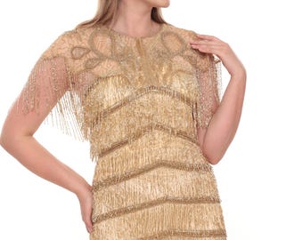 May include: A gold sleeveless dress featuring horizontal tiers of beaded fringe throughout the body. The bodice includes a sheer overlay with intricate gold beadwork in a swirling pattern and short, beaded fringe sleeves that drape over the shoulders.