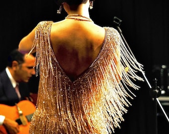 Könnte beinhalten: Eine Frau in einem goldfarbenen Kleid mit Fransen und tiefem Rückenausschnitt, von hinten fotografiert. Sie trägt eine Perlenkette und Ohrringe. Im Hintergrund spielt ein Mann im Anzug Gitarre, ein Mikrofonständer ist zu sehen.