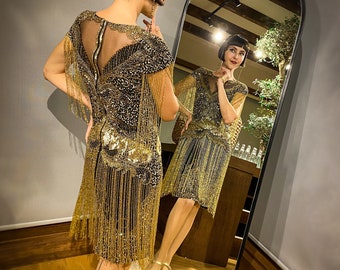 May include: A woman in a gold and black beaded flapper dress with fringe. She is standing in front of a mirror and looking at her reflection.