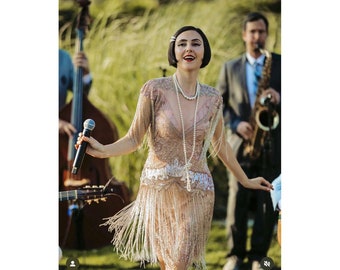 May include: A woman in a beaded flapper dress with fringe sings into a microphone. She is wearing a pearl necklace and has a bob haircut. She is performing with a band that includes a saxophone player and a double bass player.