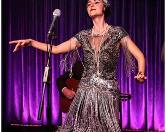 May include: A woman wearing a silver sequined flapper dress with fringe, performing on stage with a microphone. The dress has a beaded neckline and a pearl necklace.